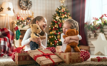 Two young girls hugging teddy bears and surrounded by Christmas gifts
