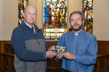 Picture of Bill Henderson and Vicar of Rushen, Reverend Joe Heaton standing in front of a stain glass window inside a church holding the new coin featuring King Charles III