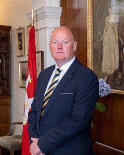 Image of Rob Callister MHK standing next to the Manx flag, in front of a painting of Queen Elizabeth II