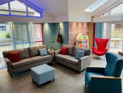 Sitting area in the Hospital Youth Service & the Bridge the Gap Pod, which has two grey sofas next to each other. There is also a blue chair next to the sofas and an orange chair behind them next to a jukebox, which has a light up rainbow ring around it.